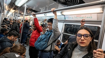 Pasajeros con abrigo dentro de un vagón de metro, algunos de pie con las manos en las barras superiores y otros sentados.