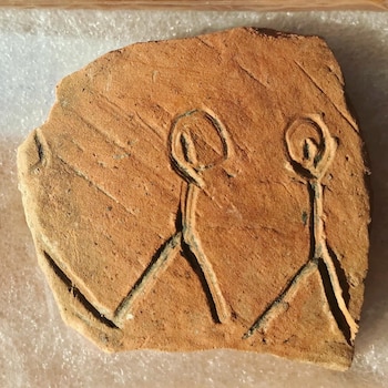 Fragmento irregular de cerámica de color terracota con dos figuras grabadas de cabeza redonda y extremidades lineales, y marcas diagonales