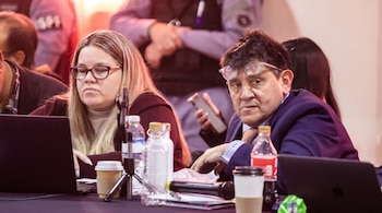 Primer plano de una mujer de gafas usando una laptop y un hombre con gafas en la frente mirando al frente, sentados en una mesa con botellas y tazas.