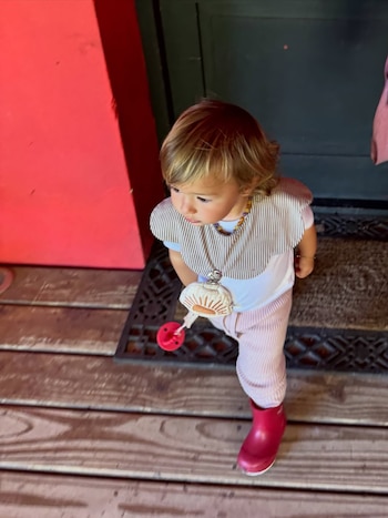 Un niño pequeño de cabello rubio claro, con camiseta a rayas, pantalones rosas, botas rojas y un collar con dije de concha y chupete, de pie en un porche de madera