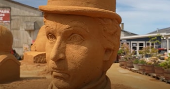 Primer plano de una escultura de arena de la cabeza de un hombre con sombrero de bombín, con facciones detalladas y expresiones serias, bajo luz natural