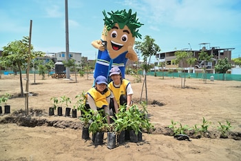 Dos jóvenes con chaleco amarillo y una mascota con disfraz de árbol posan junto a varios plantones en un terreno de tierra