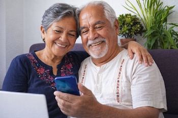 Dos adultos mayores mexicanos sonríen mientras el hombre sostiene un teléfono móvil azul. La mujer tiene su brazo sobre su hombro. Un portátil y plantas se ven en el fondo.