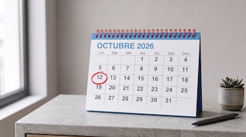 Calendario de mesa abierto en octubre de 2026 con el día 12 encerrado en un círculo rojo, junto a una maceta pequeña y un bolígrafo.