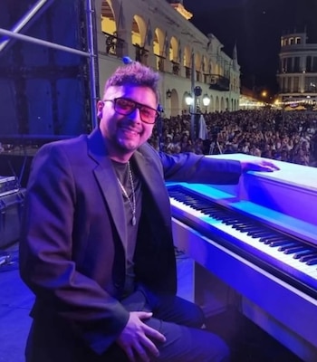 Un hombre con anteojos y saco oscuro sonríe frente a un piano blanco en un escenario nocturno con multitud