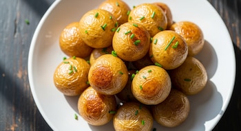 Vista superior de un plato blanco lleno de papas noisette redondas y doradas, espolvoreadas con pequeños trozos de cebollino verde, sobre una mesa oscura.