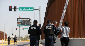 Dos policías y dos civiles, un hombre y una mujer, están junto a una escalera metálica apoyada en el muro fronterizo. Letreros de Vía Internacional y Colonia Castilla son visibles.