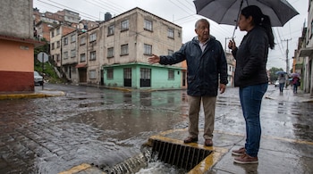Un hombre y una mujer bajo un paraguas en una calle adoquinada mojada, donde el agua corre hacia un drenaje. Al fondo, edificios residenciales en una ladera.