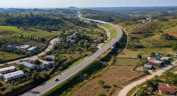 Vista aérea de una carretera renovada con un puente, serpenteando entre colinas verdes. Se observan casas dispersas, parcelas agrícolas y caminos secundarios.