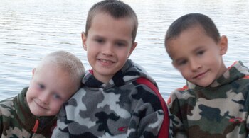 Tres niños vestidos con sudaderas de camuflaje sonríen frente al agua con la zona de los ojos desenfocada.