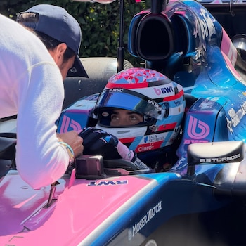 Un hombre con casco y traje de competición dentro de un monoplaza de Fórmula 3 rosa y azul. Otro hombre con gorra y suéter blanco se inclina sobre el auto