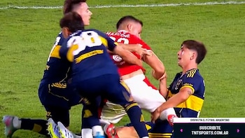 Vista cercana de tres futbolistas en el suelo y de pie, agarrándose unos a otros en el césped de un estadio. Uno lleva camiseta roja, dos azul y amarillo