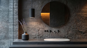 Un baño moderno con paredes de piedra oscura y textura. Incluye un espejo redondo, un lavabo blanco ovalado y grifos negros mate. Un jarrón negro con flores secas.