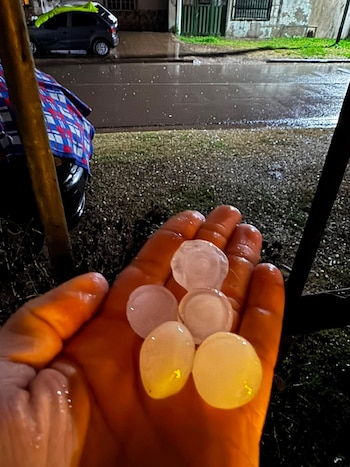 Una mano humana adulta sujeta cinco piezas de granizo ovaladas y transparentes. Al fondo se distingue una calle mojada y un coche cubierto con una lona