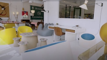 Interior moderno y luminoso con paredes blancas, un lavabo cilíndrico blanco con interior azul, mesa, sillas amarillas, un escritorio blanco y una silla de madera