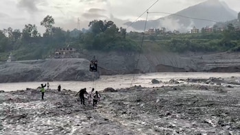 Una cesta metálica suspendida de un cable cruza un río embarrado hacia una orilla rocosa con varias personas