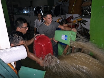 Hombres y una mujer en un interior inundado con agua turbia, usando baldes rojos y verdes para arrojar el líquido fuera del encuadre