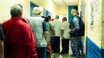 Pasillo de un edificio público con paredes amarillas y azulejos azules. Hombres en fila y un guardia uniformado con bandera de Guatemala en el hombro, mochila y bastón