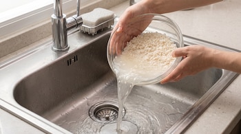 Dos manos sostienen un tazón de vidrio con arroz y vierten agua blanquecina en un fregadero de metal.