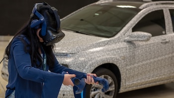 Una persona con cabello oscuro y blusa azul oscuro usa un visor de realidad virtual y dos controladores, frente a un coche camuflado en un centro de desarrollo