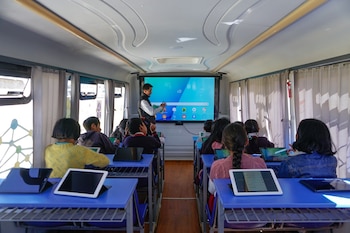 Interior de un bus con un profesor de pie frente a una pantalla grande. Alumnos están sentados en pupitres azules usando tabletas o laptops