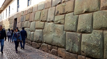 Pared de mampostería inca con grandes bloques de piedra de tonos tierra y gris, algunos con musgo, en una calle empedrada con personas y un edificio blanco al fondo.