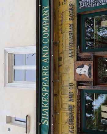 Fachada de una librería con toldo verde que dice "Shakespeare and Company". Cartel con "Antiquarian Books" y un retrato de William Shakespeare. Ventanas verdes
