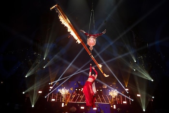 Un hombre y una mujer vestidos de rojo realizan acrobacias aéreas suspendidos de un tronco en llamas sobre un escenario con luces