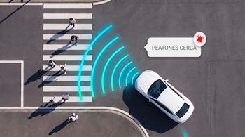 Vista aérea de un automóvil blanco ante un paso de peatones con transeúntes y un aviso digital con el texto peatones cerca.