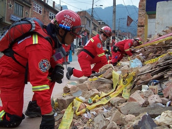 Tres rescatistas con uniformes rojos, cascos y mochilas, de rodillas entre escombros. Cinta amarilla de seguridad en el suelo. Edificios y montañas al fondo