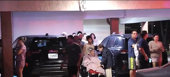 Un grupo de personas y una persona en silla de ruedas permanecen de noche junto a dos camionetas bajo un estacionamiento techado
