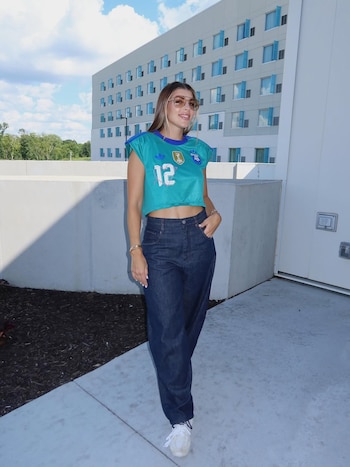 Mujer de pie con camiseta corta turquesa con número doce, escudo FIFA y logo AFA, jeans oscuros, zapatillas blancas, y gafas de sol, ante un edificio