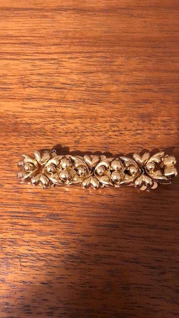 Primer plano de un broche dorado alargado con un patrón floral y esferas brillantes, apoyado sobre una mesa de madera oscura con vetas
