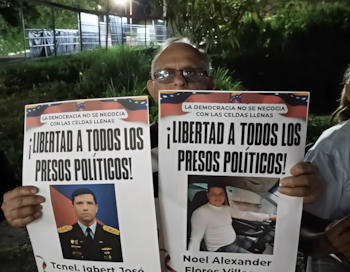Un hombre con anteojos sostiene dos carteles con retratos y el texto Libertad a todos los presos políticos junto a la bandera de Venezuela