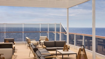 Terraza de crucero con cómodos sofás, sillas y mesas de centro bajo una pérgola, con barandillas de cristal y el mar abierto de fondo