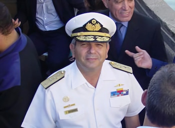 Hombre con uniforme naval blanco, gorra de plato con emblema dorado, hombreras doradas, medallas y barras en el pecho. Fondo con otras personas