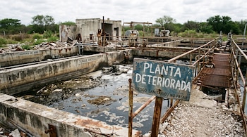 Un cartel de madera con el texto Planta deteriorada junto a piletas de concreto con agua oscura y pasarelas metálicas oxidadas.