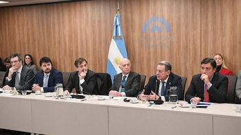 Seis hombres sentados en una mesa de conferencia con micrófonos, vasos, tazas y documentos. Al fondo, una pared de madera, una bandera argentina y un logo azul de 'Diputados'