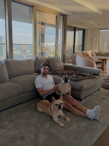 Un hombre sentado en el suelo con dos perros bulldog francés. Detrás hay un sofá y una gran ventana con vista a edificios y el cielo