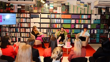 Tres mujeres en sillones frente a una audiencia en una librería. Estanterías con libros de fondo, una pantalla de TV y mesas pequeñas con libros