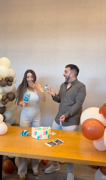 Una mujer y un hombre sostienen copas con pastel azul junto a una mesa con un pastel cortado, ecografías y globos