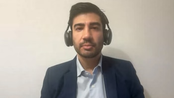 Hombre con cabello oscuro y barba, camisa clara, saco oscuro, y auriculares negros sobre un fondo claro