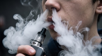 Primer plano de la boca y nariz de un hombre que exhala vapor de un vapeador gris que sostiene con la mano derecha.