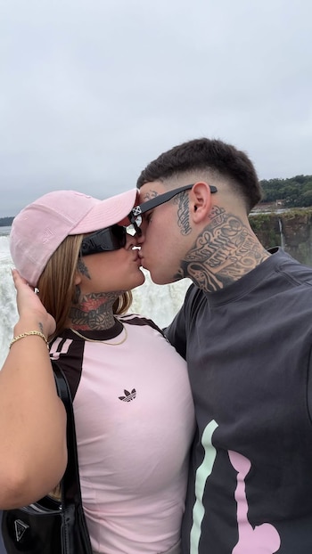Tamara Báez y su pareja se besan en un primer plano, con las Cataratas de Iguazú de fondo. Ella lleva gorra rosa y gafas de sol, él tiene tatuajes en el cuello