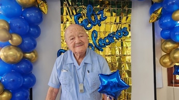 Un hombre mayor con camisa azul sonríe frente a un fondo dorado de "Feliz Cumpleaños" y globos azules y dorados, sosteniendo un globo de estrella azul