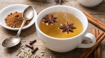 Primer plano de una taza blanca de infusión humeante con dos vainas de anís estrellado flotando, rodeada de ramas de canela, clavos y semillas de hinojo sobre madera.