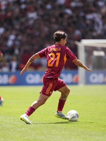 O jogador de futebol Paulo Dybala por trás, vestindo camisa e shorts cor de vinho, número 21, chutando uma bola branca no campo de futebol
