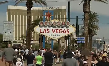 Varias personas caminan frente al cartel de Las Vegas entre palmeras y postes, con el edificio del hotel Mandalay Bay al fondo