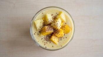 Vaso transparente con smoothie cremoso amarillo, trozos de ananá, trozos de mango y semillas de lino espolvoreadas en la superficie, fondo de madera clara.