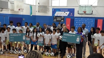 En un gimnasio, varios niños con camisetas deportivas y adultos posan sonriendo, algunos con balones de fútbol, frente a pancartas azules del Mundial 2026